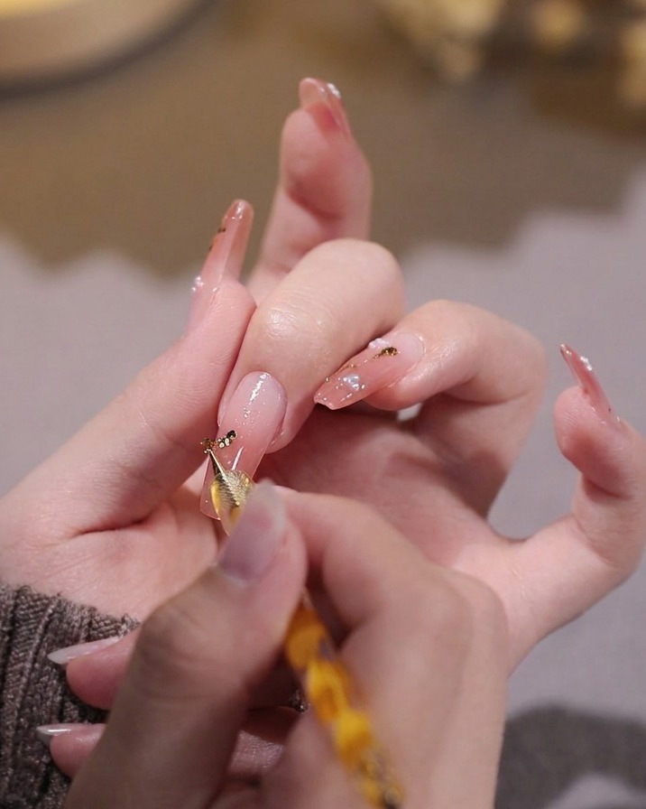 A hand using a dotting tool to place tiny gold foil flakes onto a pink gel-painted nail.