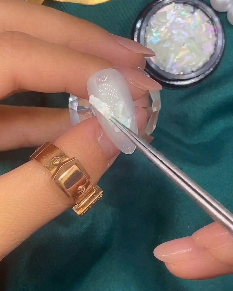 A brush applying tiny gold foil flakes onto the surface of a nail already decorated with shell pieces.