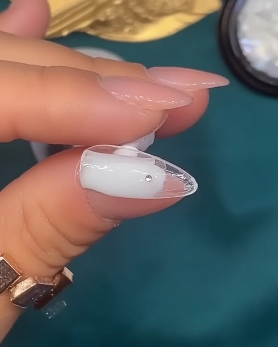 A brush applying a translucent, milky white gel polish to a natural-shaped nail.