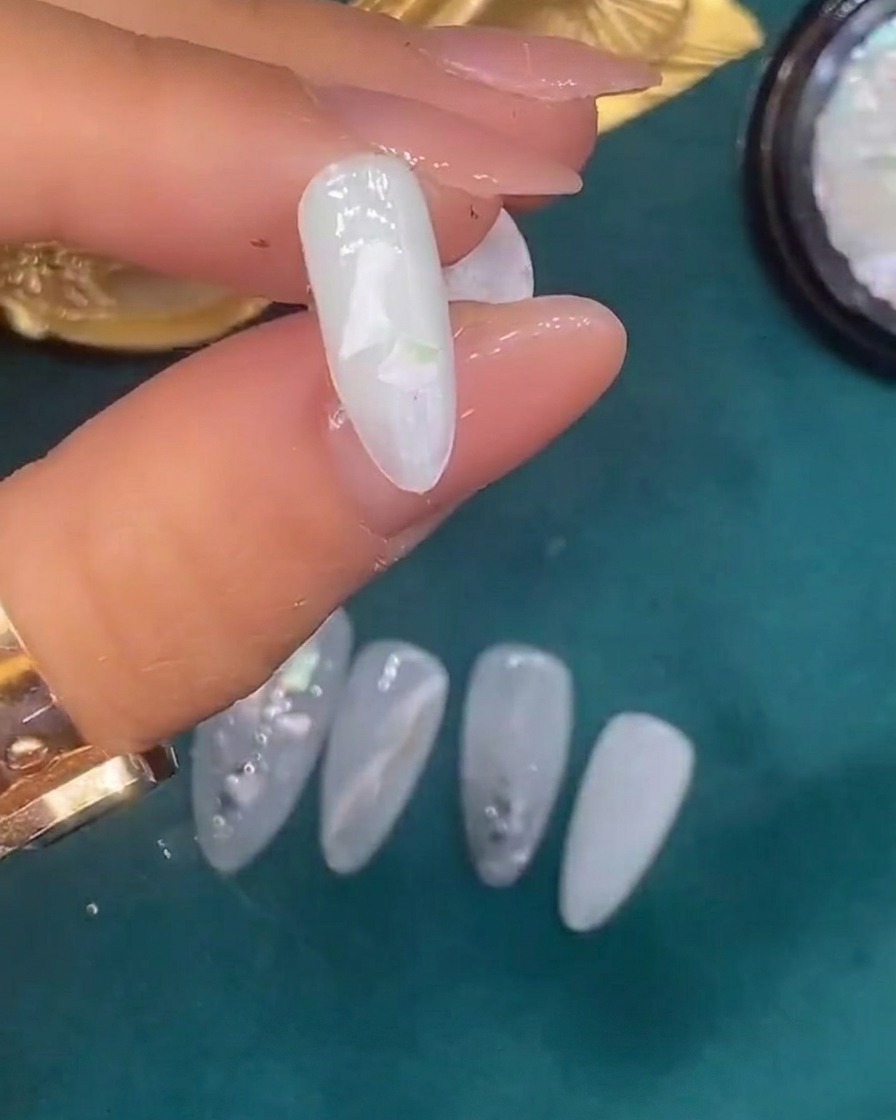 Tweezers placing decorative shell pieces onto a nail coated in wet, translucent white gel.