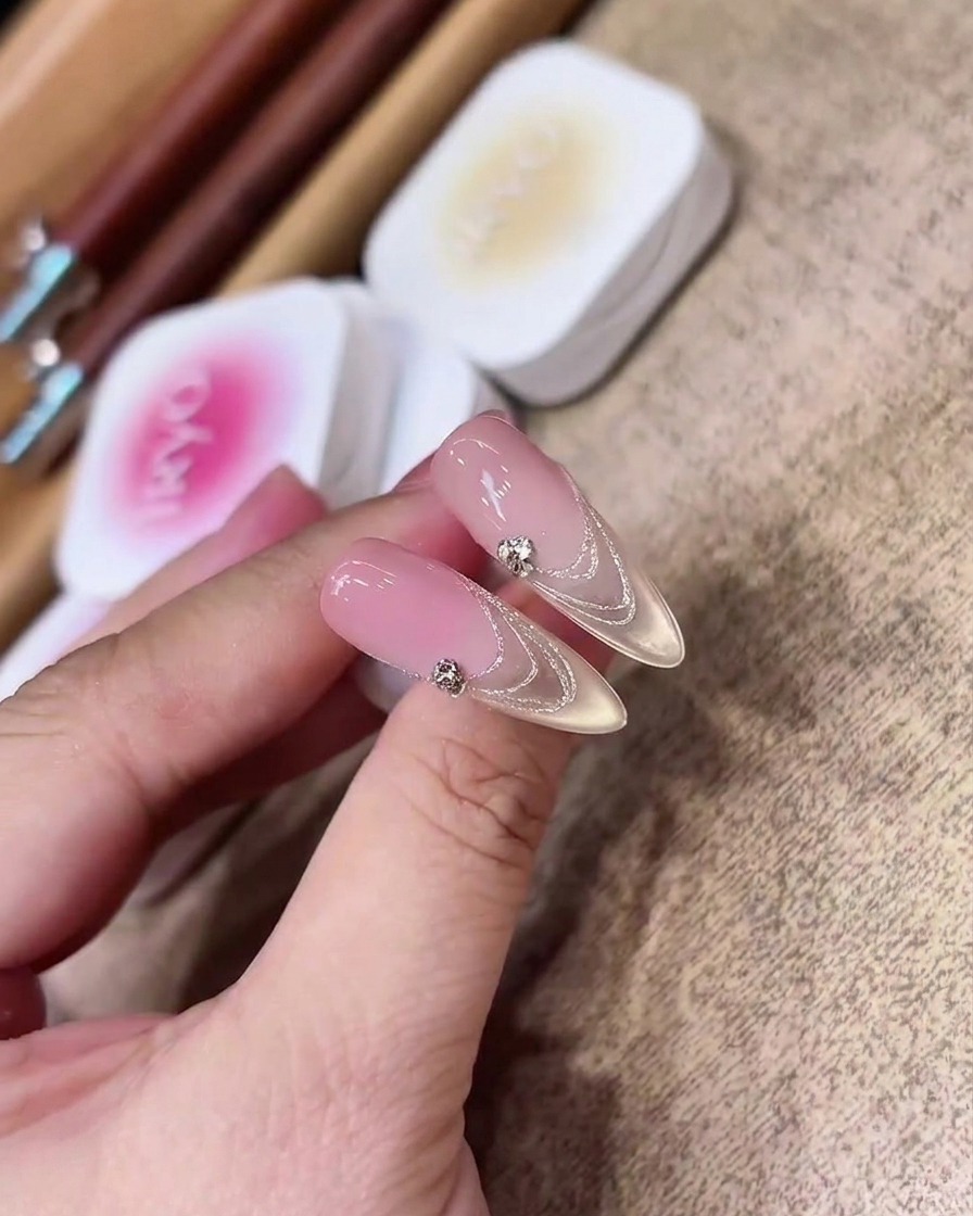 A close-up view of a flat gel brush evenly applying a sheer pinkish-nude gel onto a clear, almond-shaped nail extension.