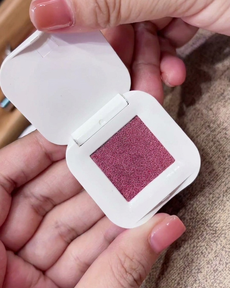 A close-up of a triangular makeup sponge pressing into a square compact of fine pinkish-purple pigment.