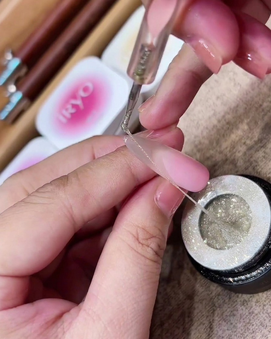 A dotting tool lifting a long, thin, stretchy strand of silver glitter spider gel from a jar.
