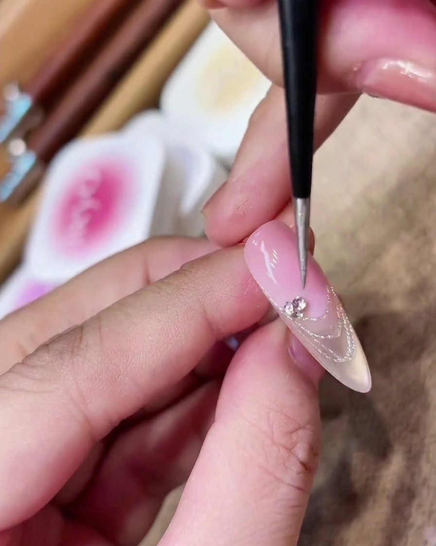 A detail brush placing a cluster of small, sparkling rhinestones onto the smile line of a French manicure.
