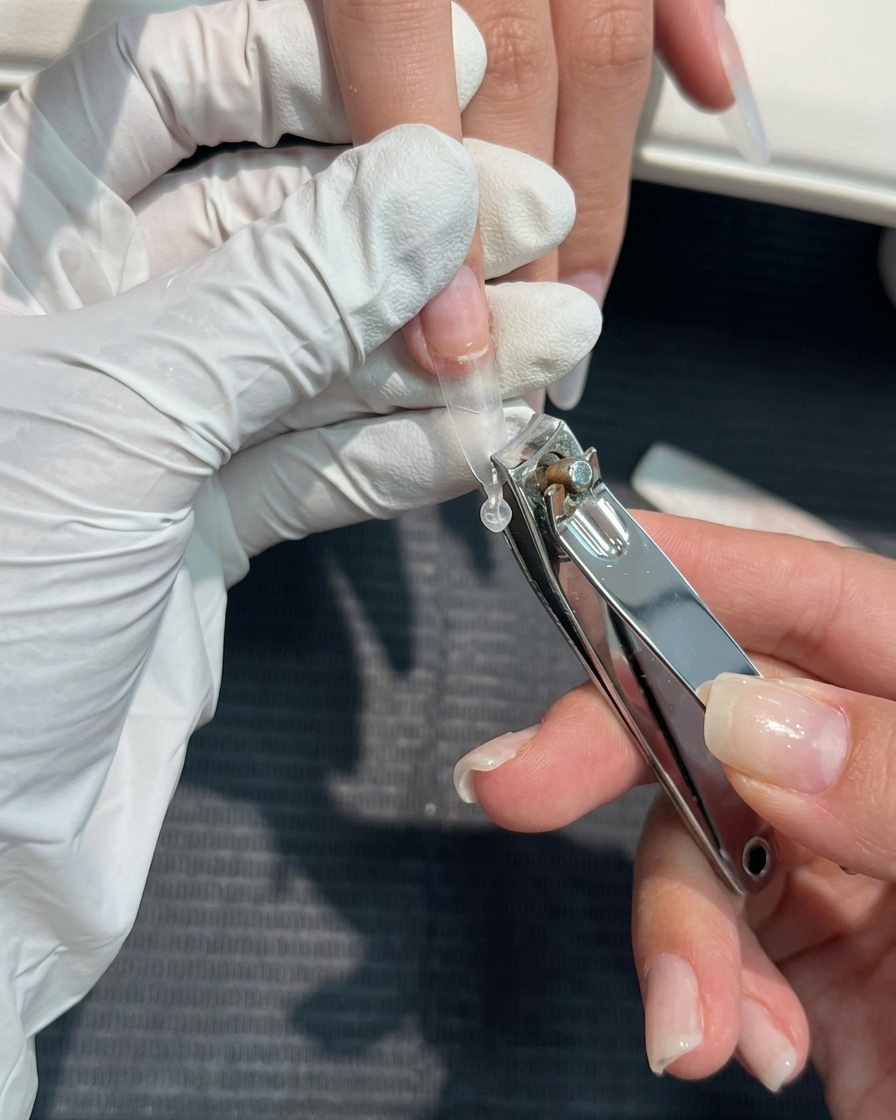 A close-up view of nail clippers being used to trim a clear plastic nail extension tip to the desired length.