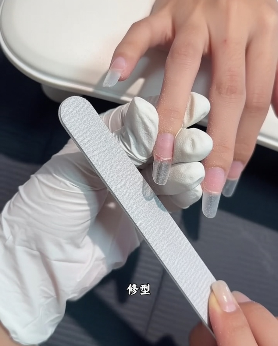 A hand holding a file to shape the edges of a clear nail extension.