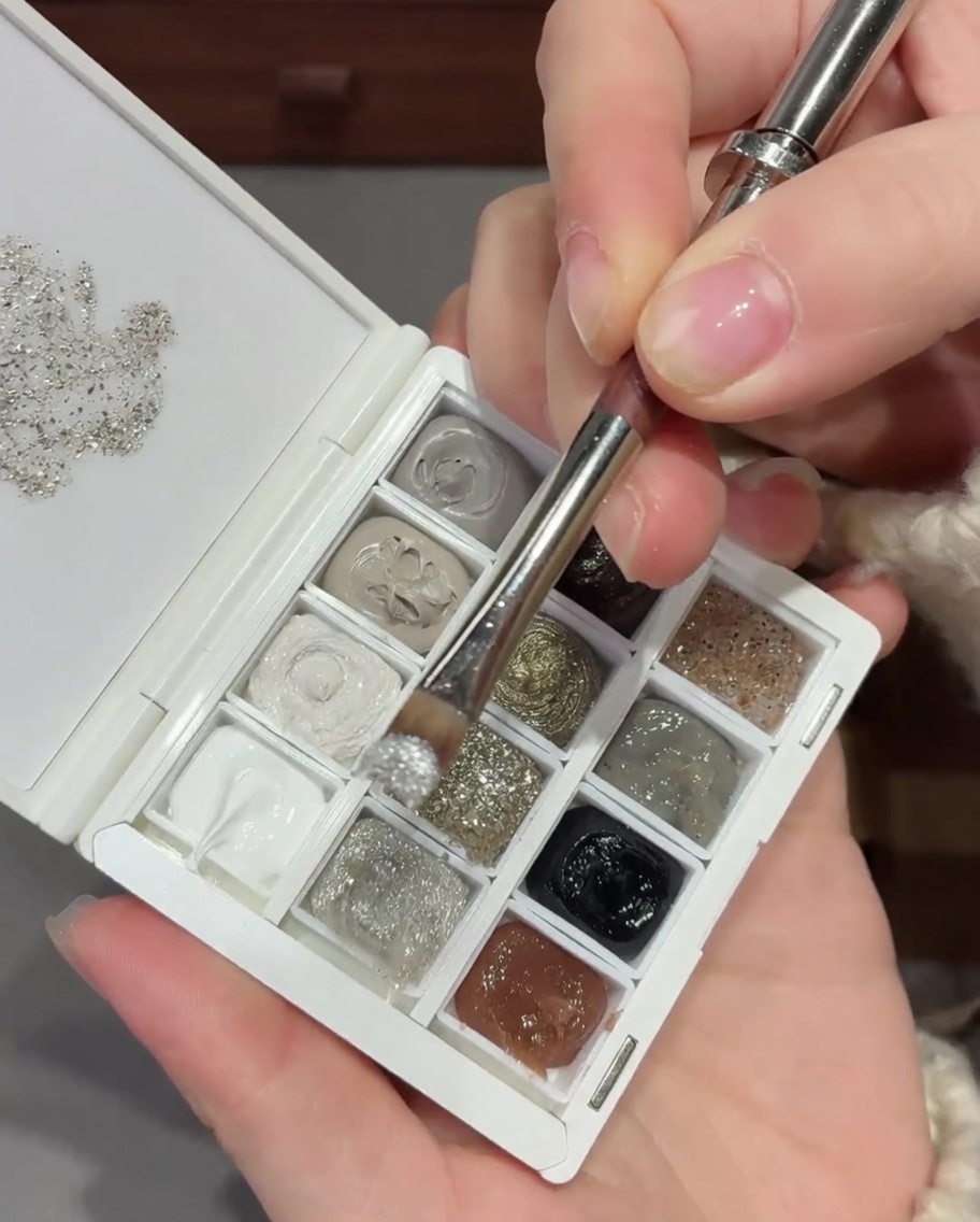 A nail brush applying a silver glitter and sequin mixture to the base of a natural nail.