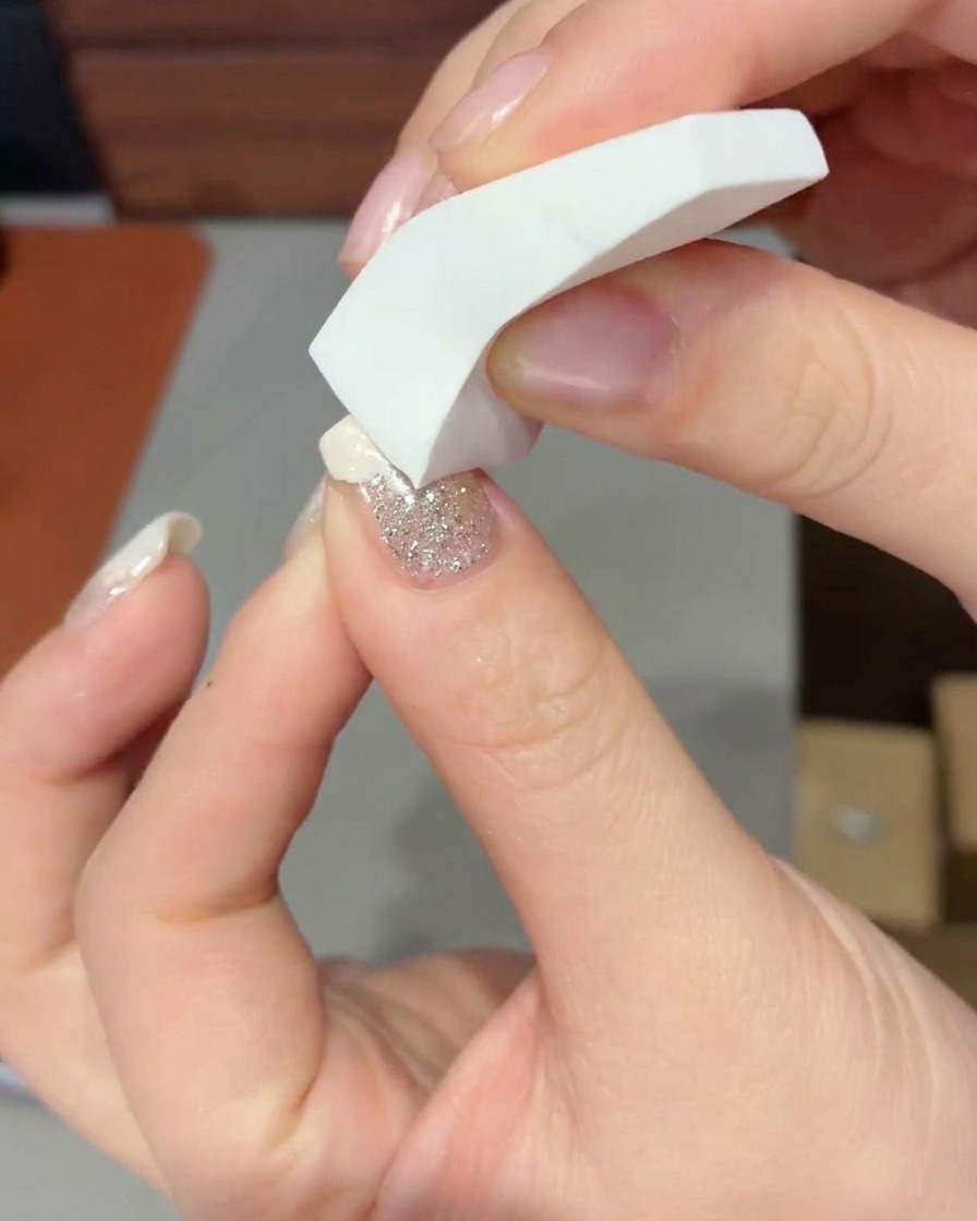 A close-up of a makeup sponge dabbing milky white gel onto the tip of a nail to form a soft gradient.