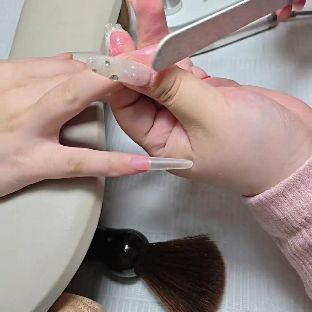 A nail technician using a hand file to blend the cuticle seam of a newly applied clear stiletto nail tip.