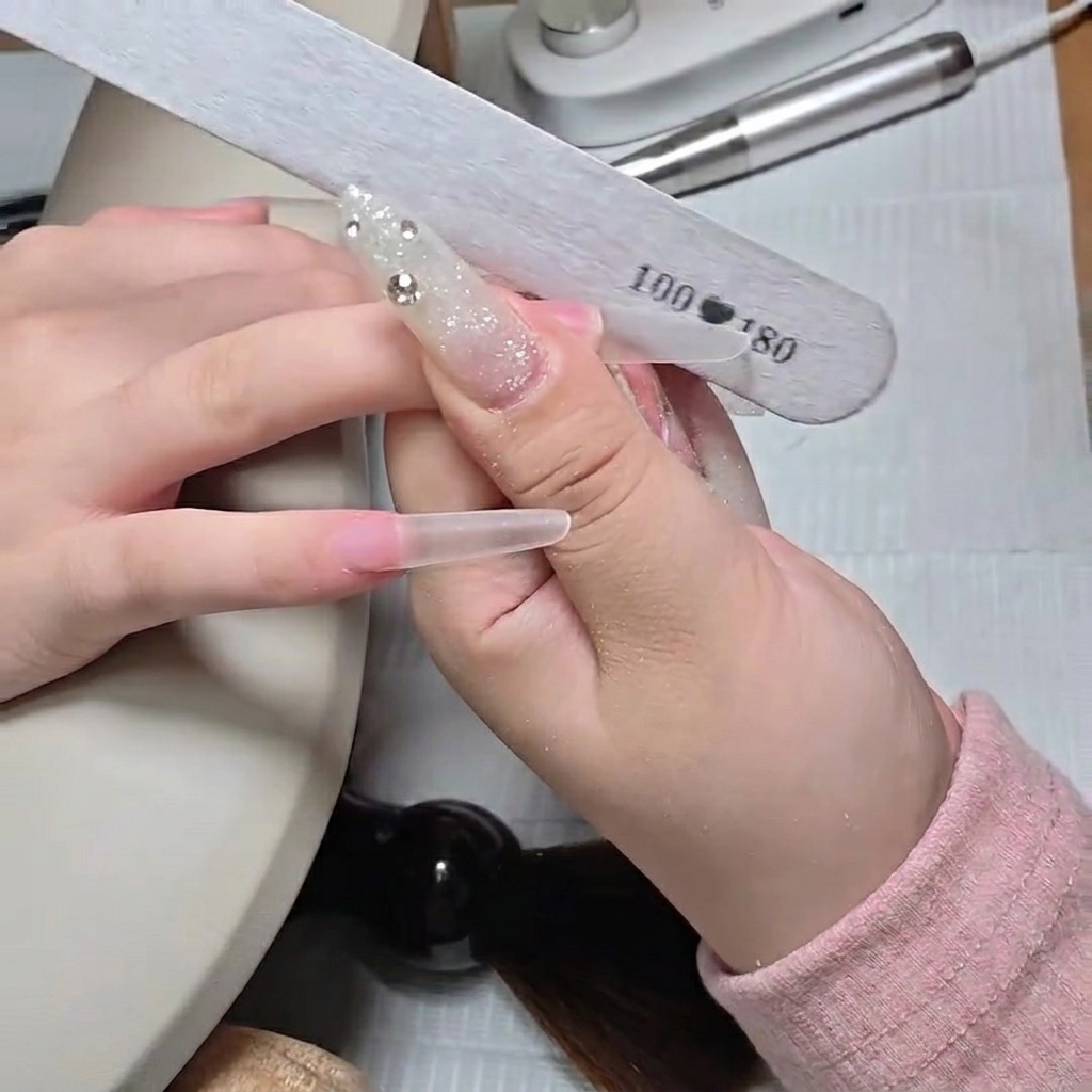 A close-up of a hand file shaping the free edge of a long clear stiletto nail tip.