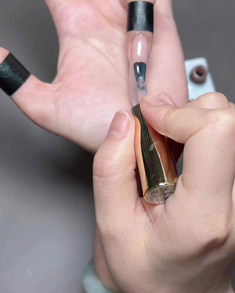 A brush applying a light, sheer brown-toned gel polish to a long natural nail.