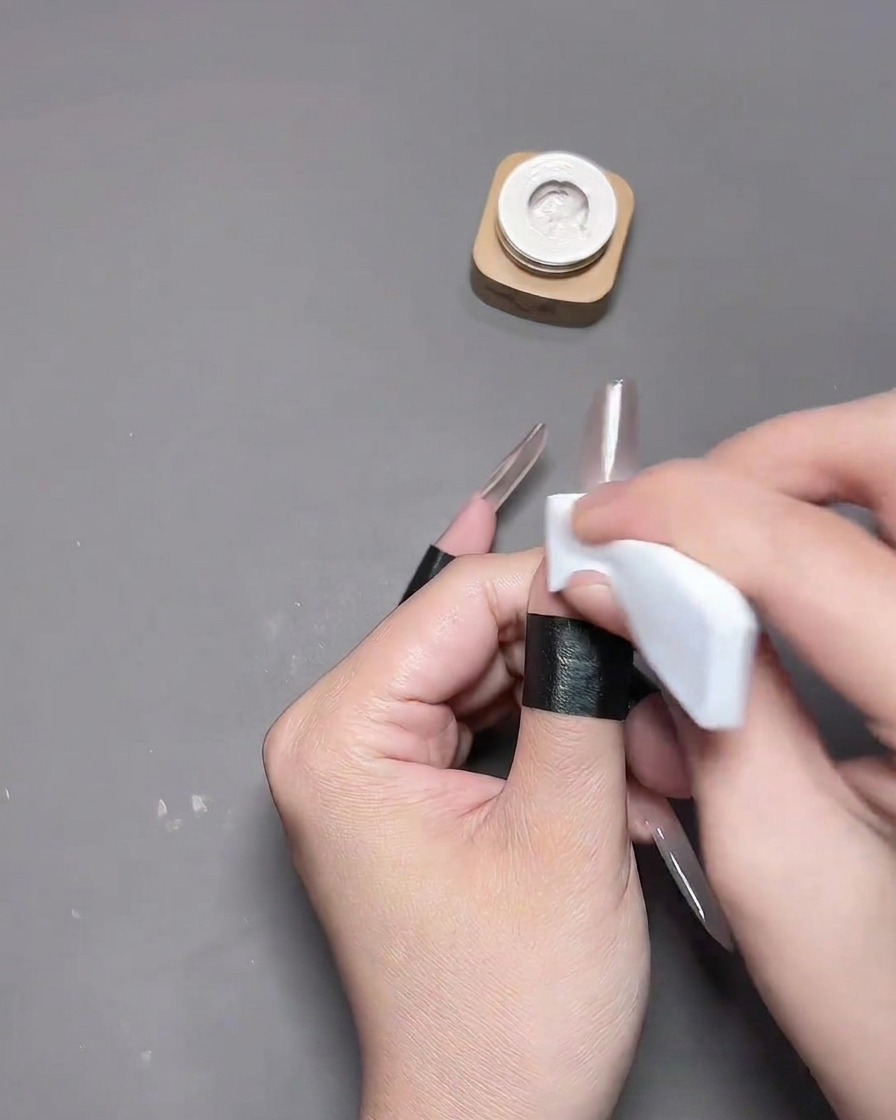 A close-up of a hand pressing a hollow heart stencil sticker onto a thumb nail with a white sponge applicator, while the surrounding skin is protected with black barrier tapecite: 1.