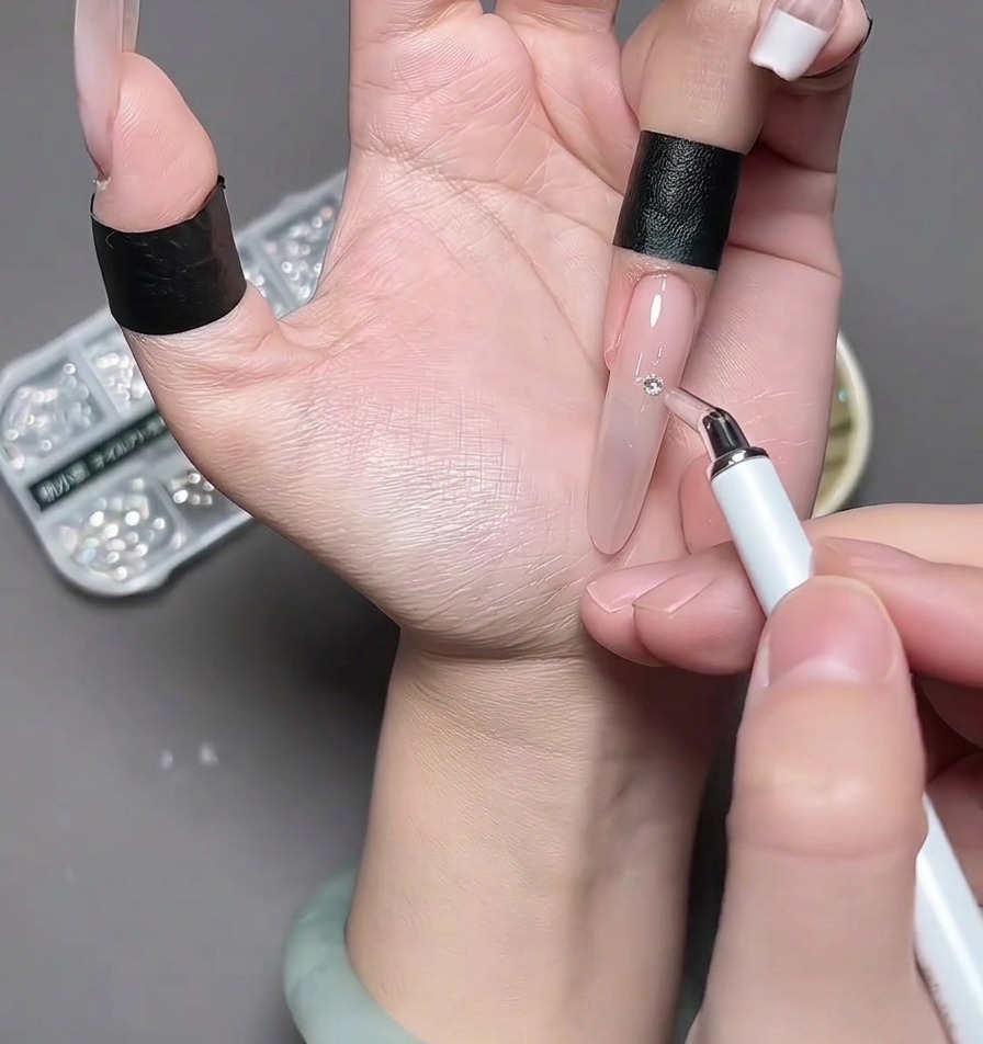 Tweezers placing a small, silver chrome heart charm onto the center of a fingernail.