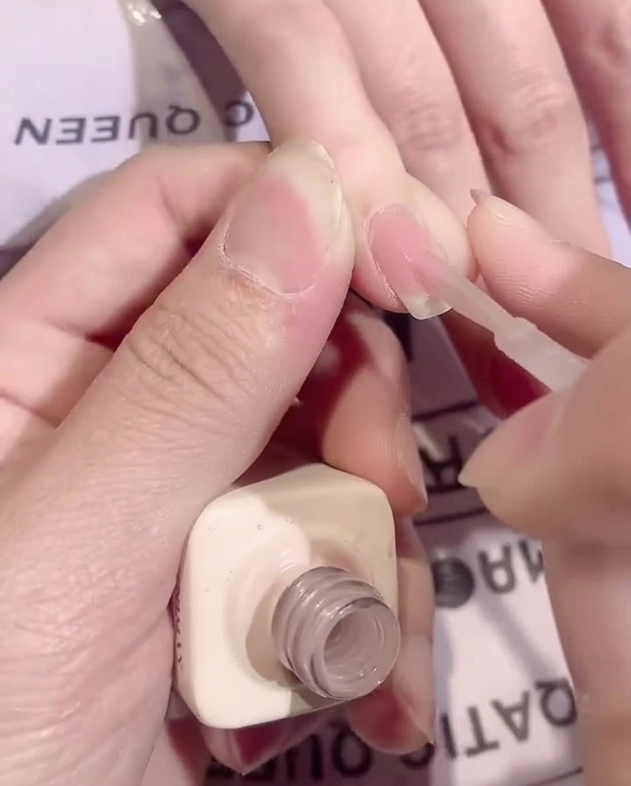 A brush applying a smooth, thin layer of sheer nude base coat gel onto a fingernail.