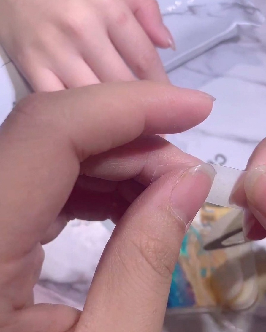 Fingers carefully placing a thin strip of clear isolation tape across a fingernail to block off the base.