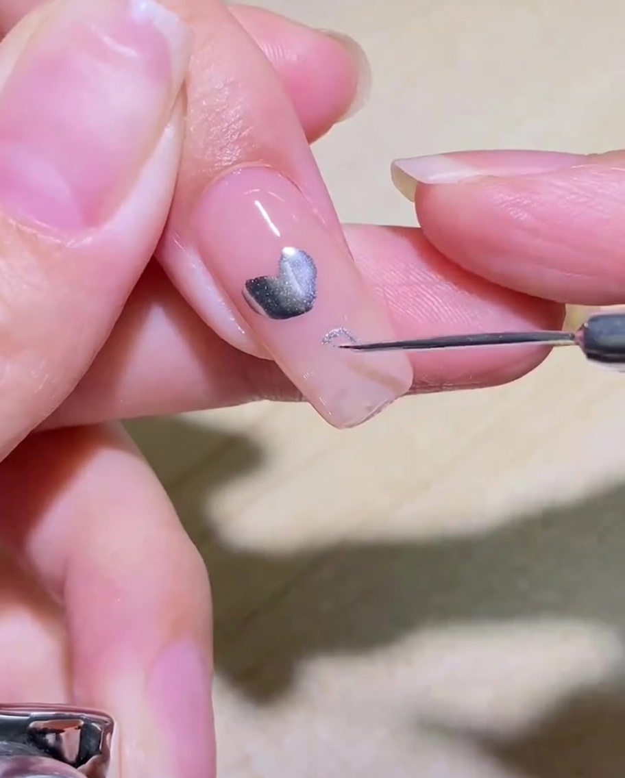 A close-up of a liner brush drawing a silver heart outline on a clear-coated nail.