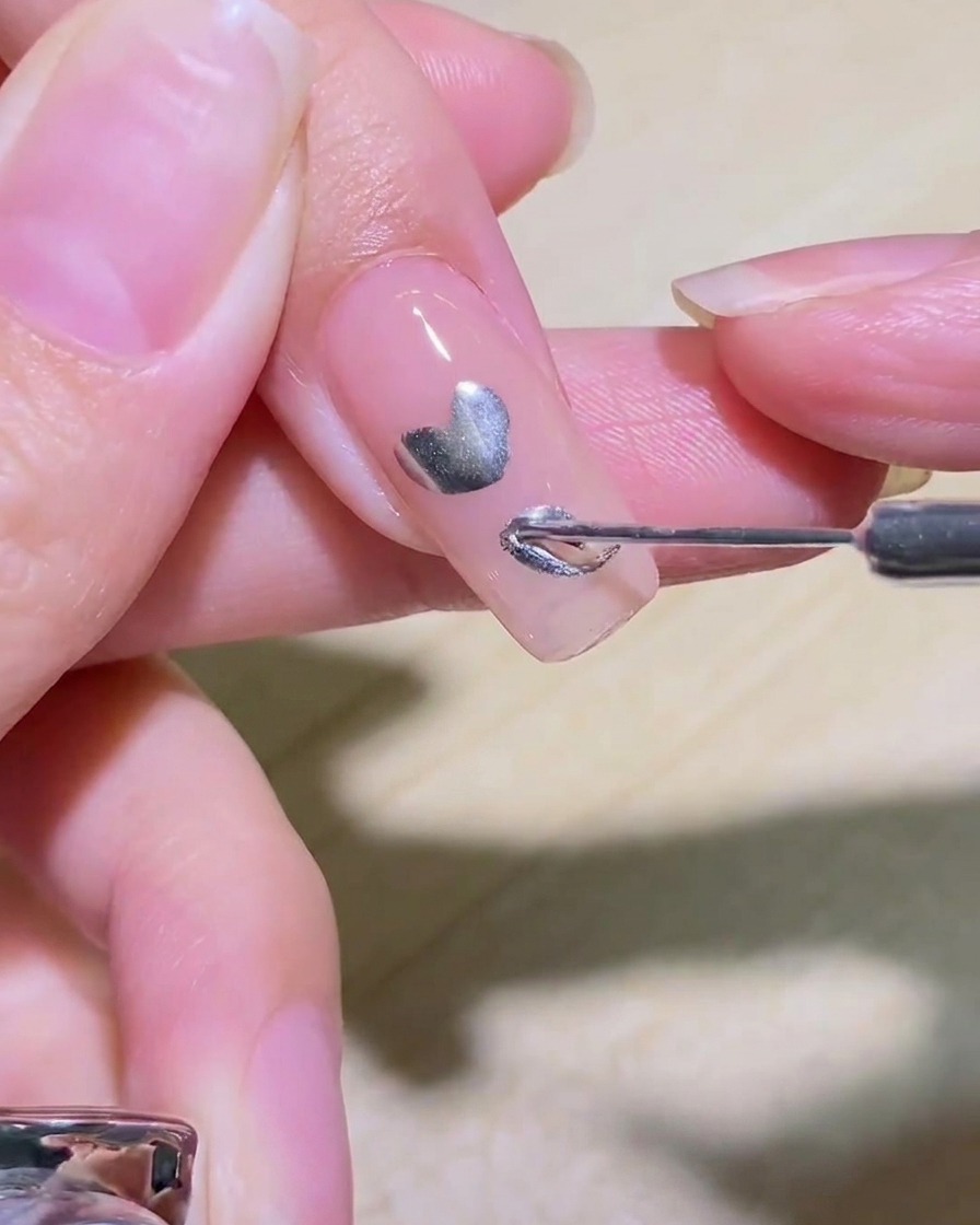 A liner brush filling in the interior of a silver heart design on a nail.