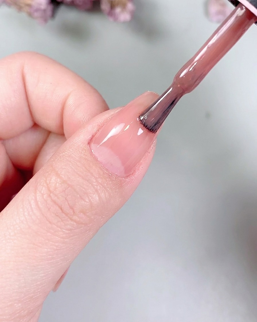 A small flat gel brush gently smoothing the nude color across the nail surface.