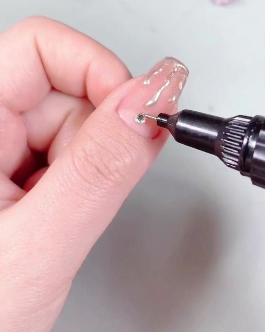 A nail art brush applying a layer of high-gloss steel top coat over a nail finished with flame art and a rhinestone.