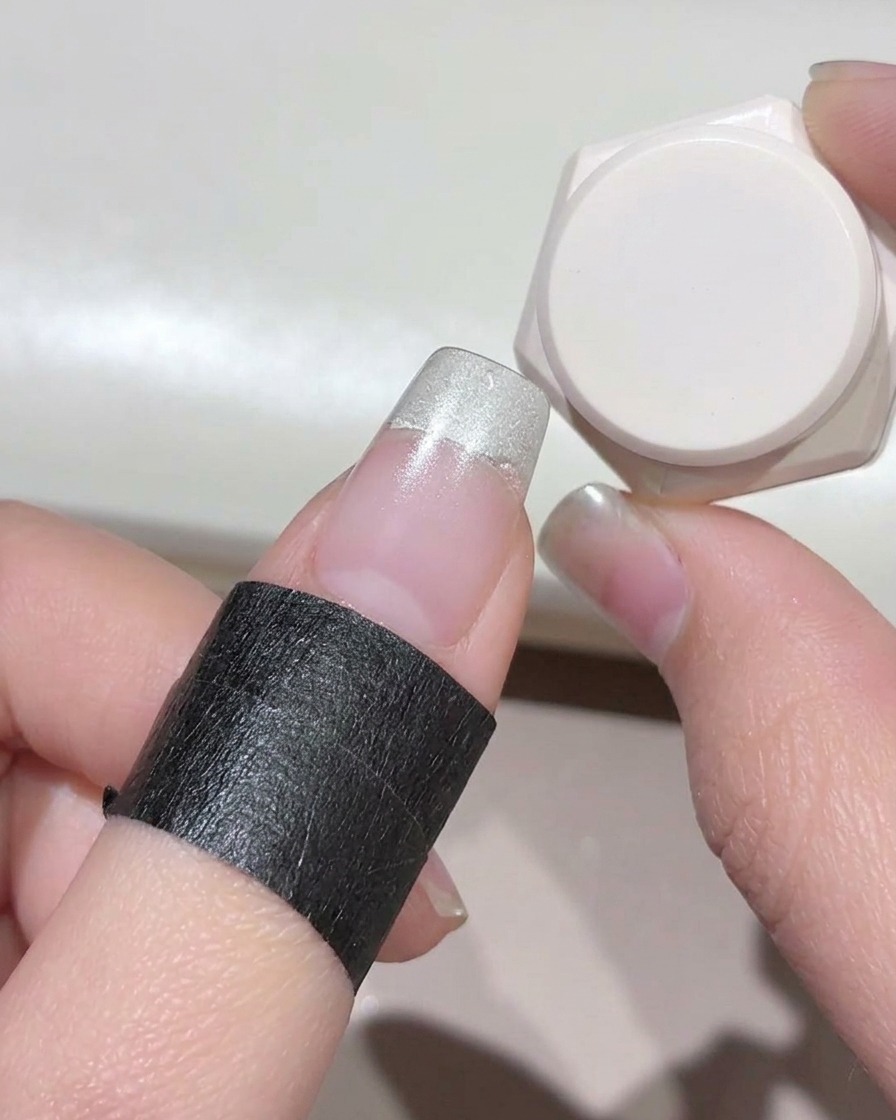 A close-up of a finger with black protective tape wrapped around the skin, showing milky white gel applied to the nail tip.