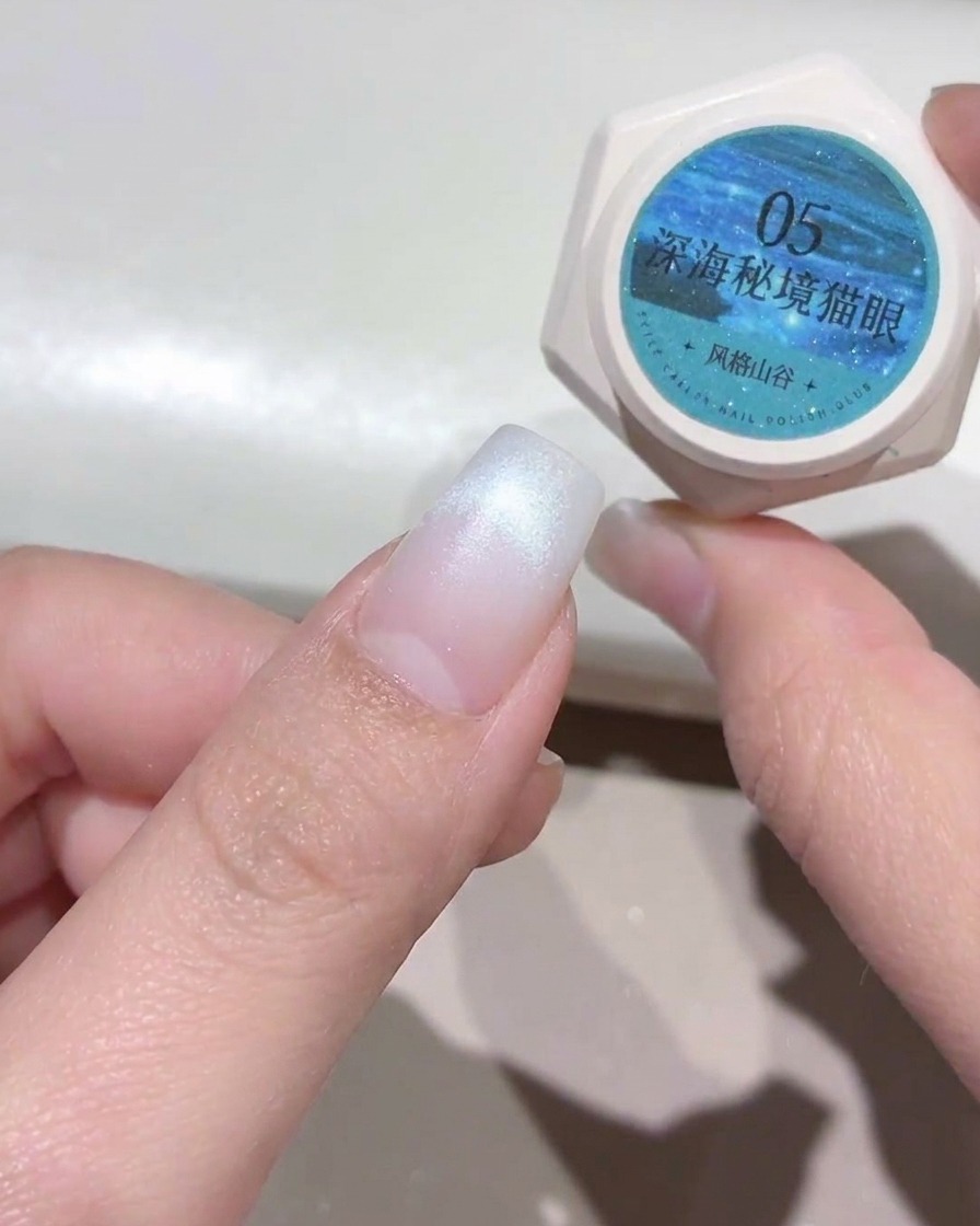 A hand holding a small pot of teal-blue cat-eye gel next to a prepared fingernail with a white tip.
