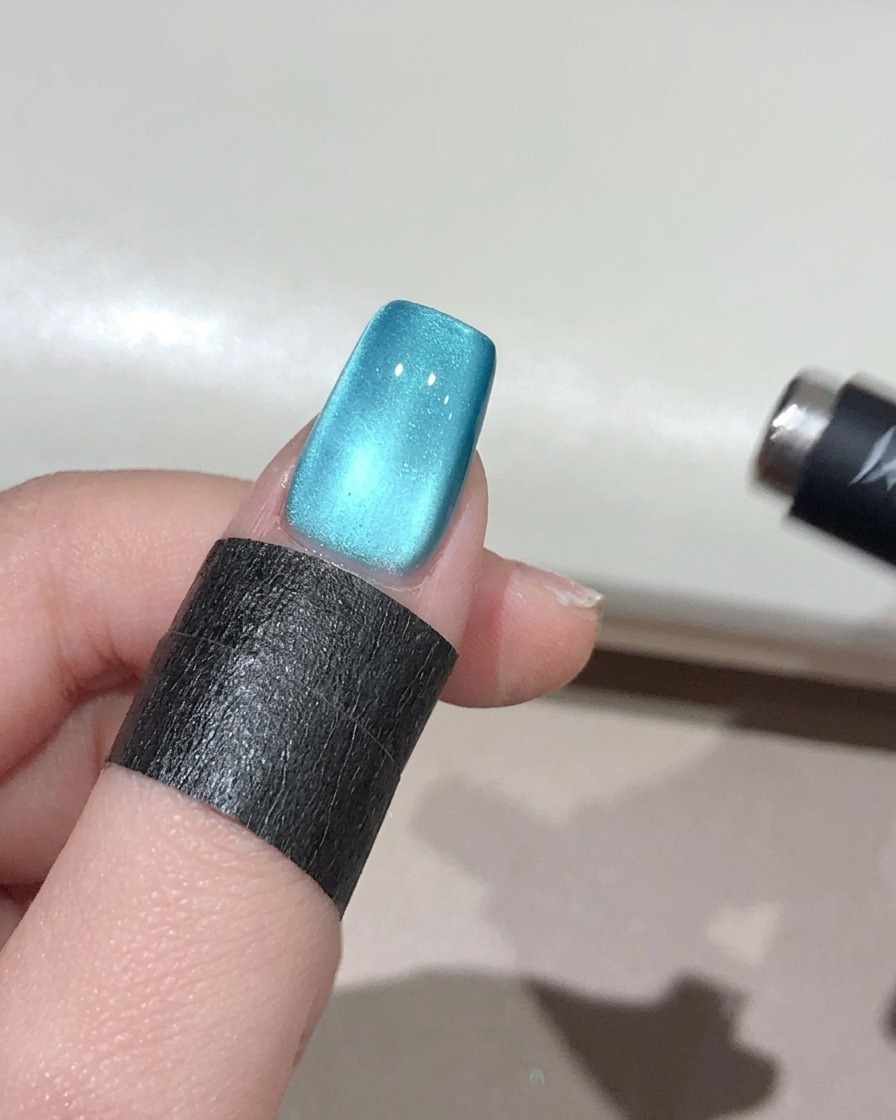 A small spherical magnet held near the edge of a teal-blue nail, concentrating the shimmer into a glowing, 3D bead effect.