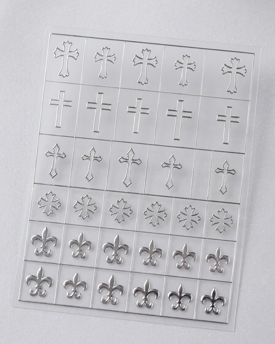 A close-up view showing fine-tipped tweezers lifting a delicate, silver-colored cross-shaped stencil sticker from a clear adhesive sheet.