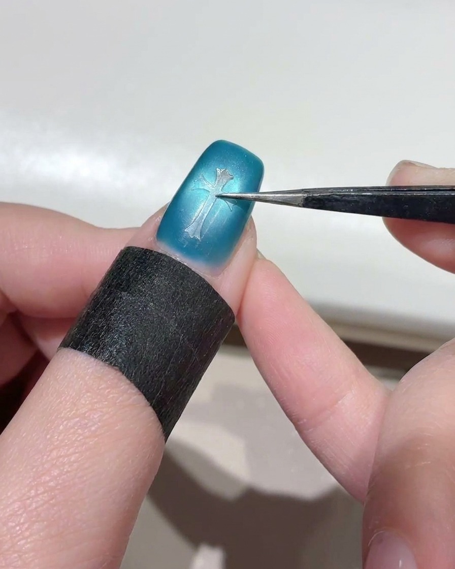 A close-up of tweezers pressing a clear cross-shaped stencil sticker onto the center of a shimmery blue cat-eye nail.