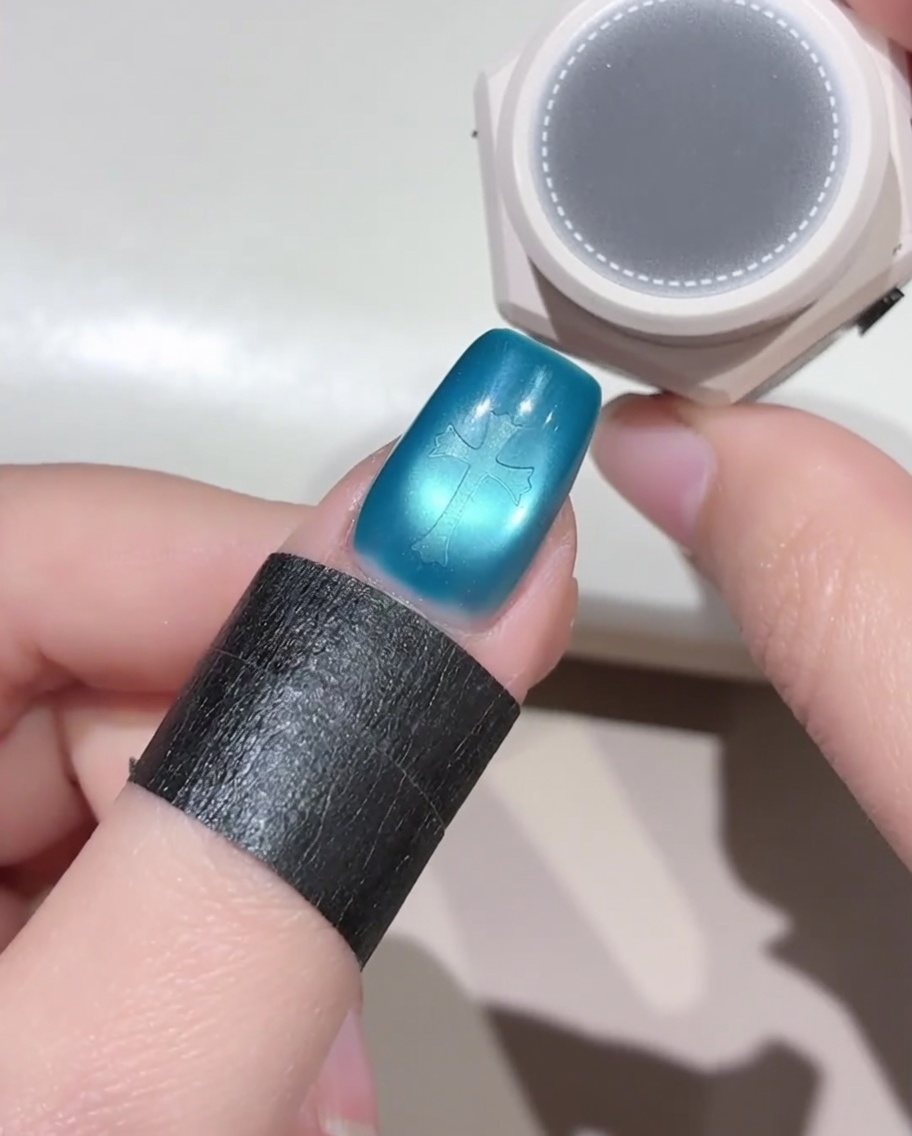 A small round sponge tool hovering over a blue fingernail that has a cross-shaped sticker on it, preparing to apply black gel polish.