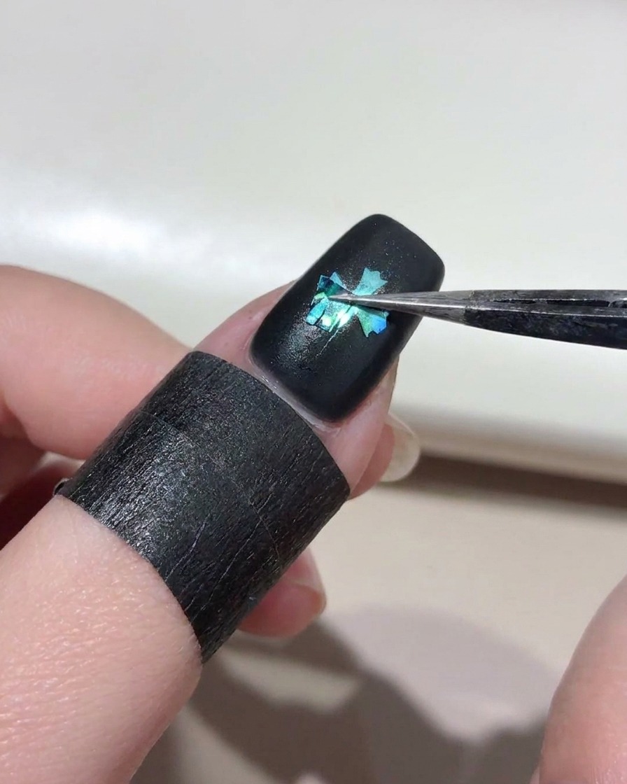 Tweezers carefully lifting a cross stencil from a black-painted nail, revealing a bright blue cat-eye cross shape underneath.