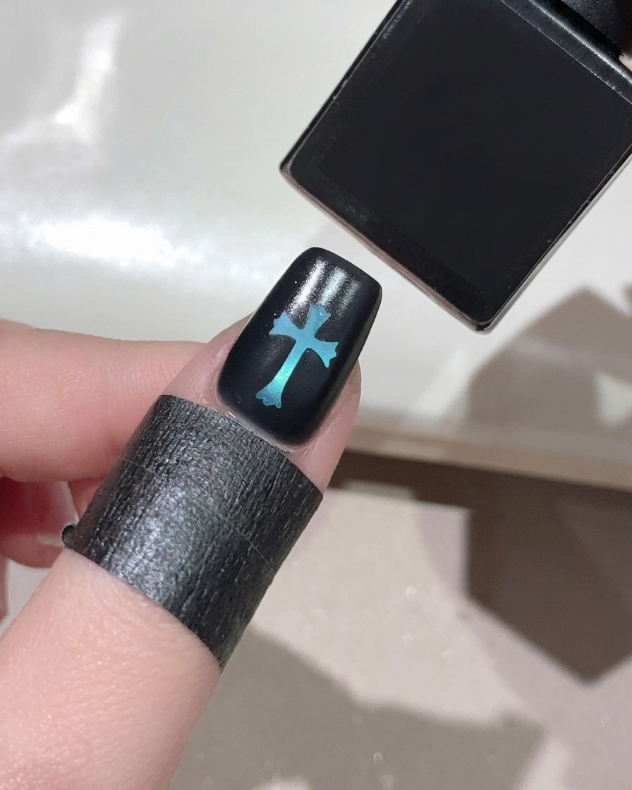 A brush applying a clear glossy top coat over a black nail with a glowing blue cross design in the center.