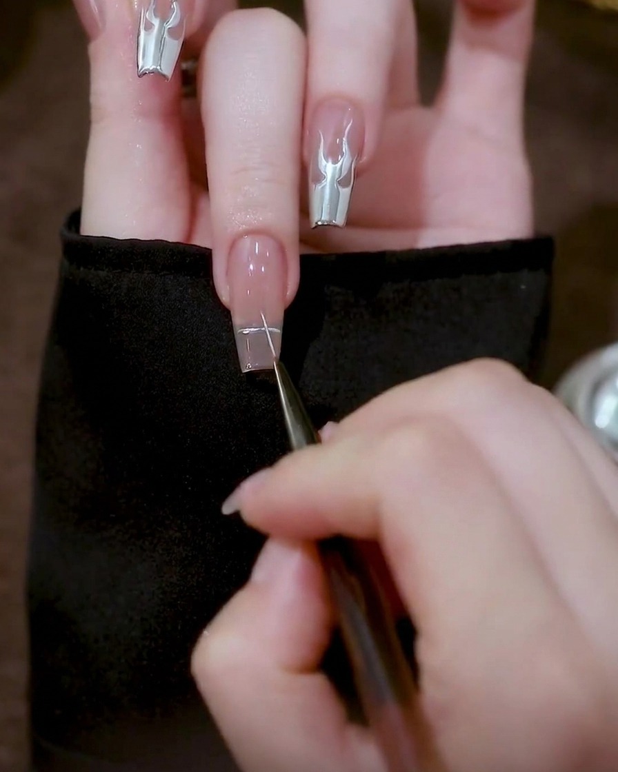 A close-up view of a fine liner brush marking a small cross with silver gel at the center of a clear nail tip.