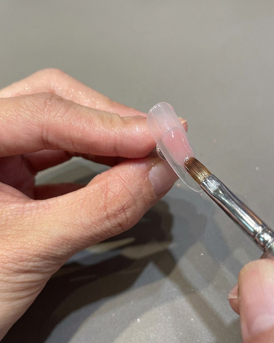 A professional nail brush applying a sheer, translucent pink gel polish to a prepared nail surface.