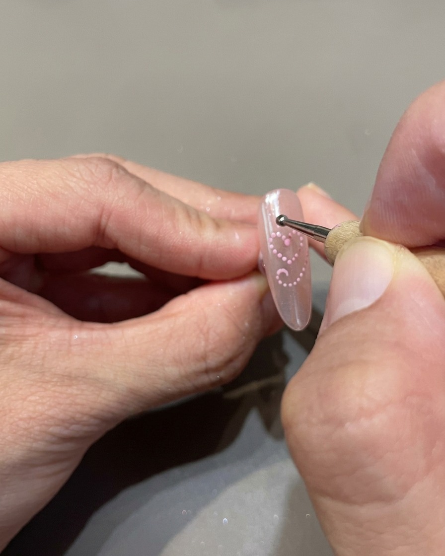 A dotting tool placing small, uniform dots of pink cat-eye gel to outline a heart on the nail.