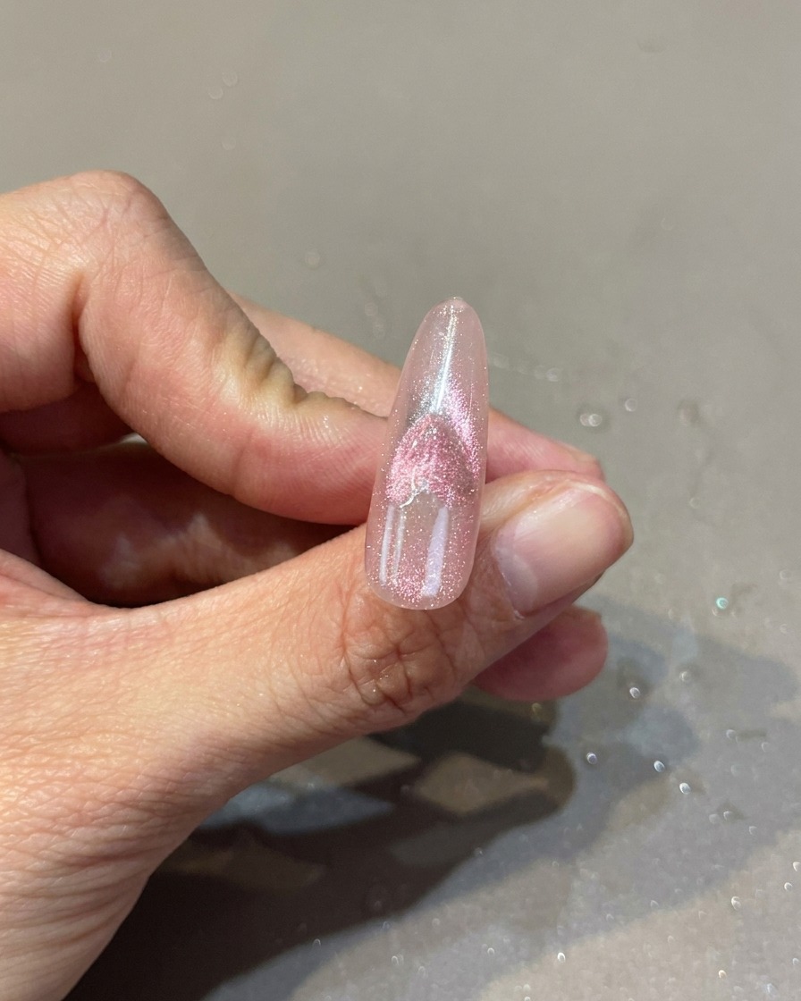 A close-up of a pink nail with a heart-shaped cat-eye design, showing a magnet being used to manipulate the shimmer effect.