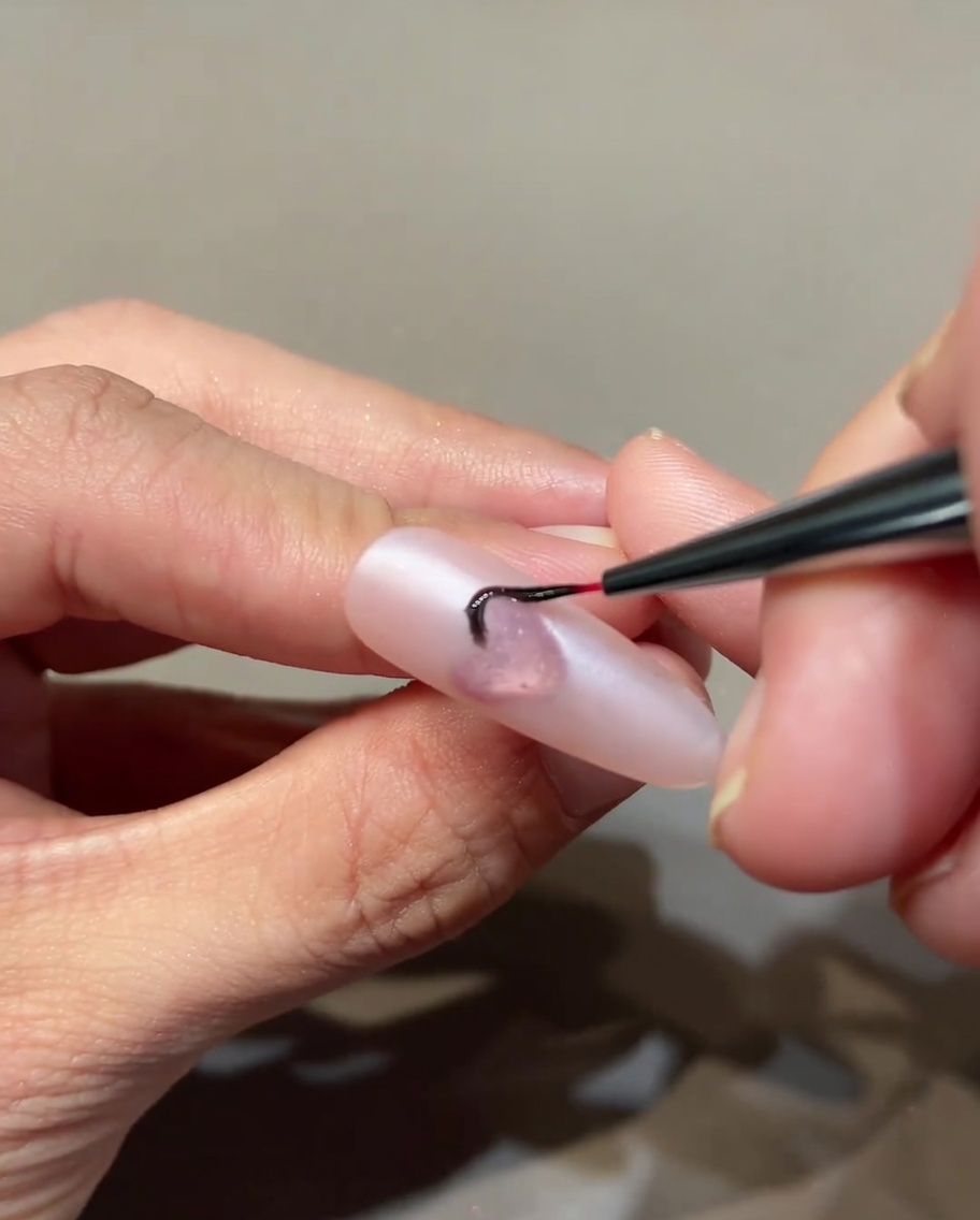 A thin nail art brush drawing fine black flame details on a pink-based nail around a central heart.