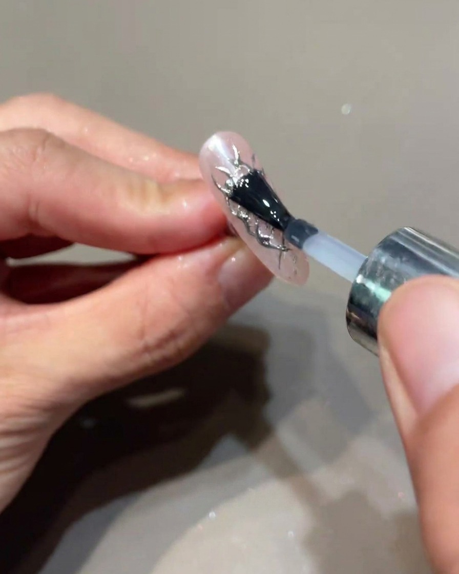 A brush applying a final layer of clear top coat over a nail decorated with silver flame accents.