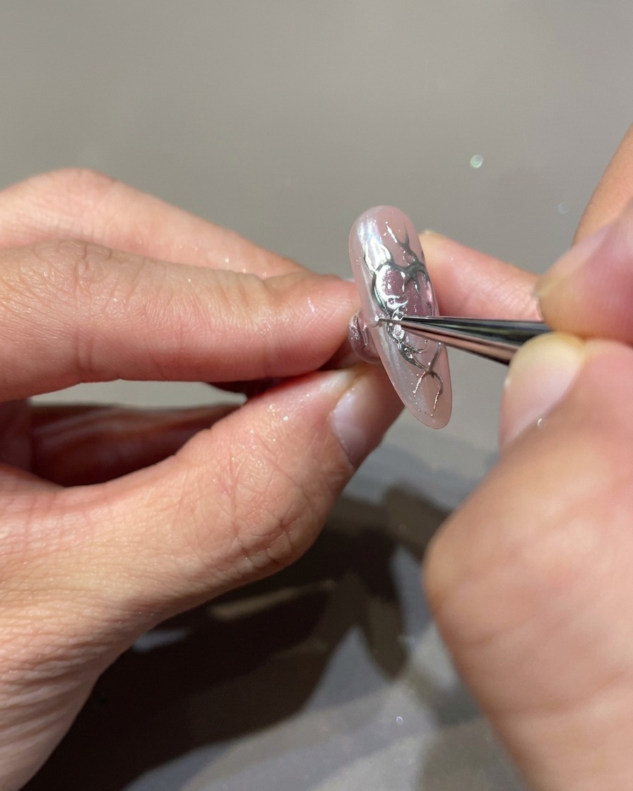 A close-up of a nail being decorated with a fine brush applying clear 3D builder gel over black flame outlines.
