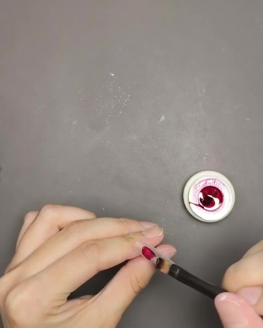 A close-up of a brush applying deep red cat eye gel polish onto a nail tip.