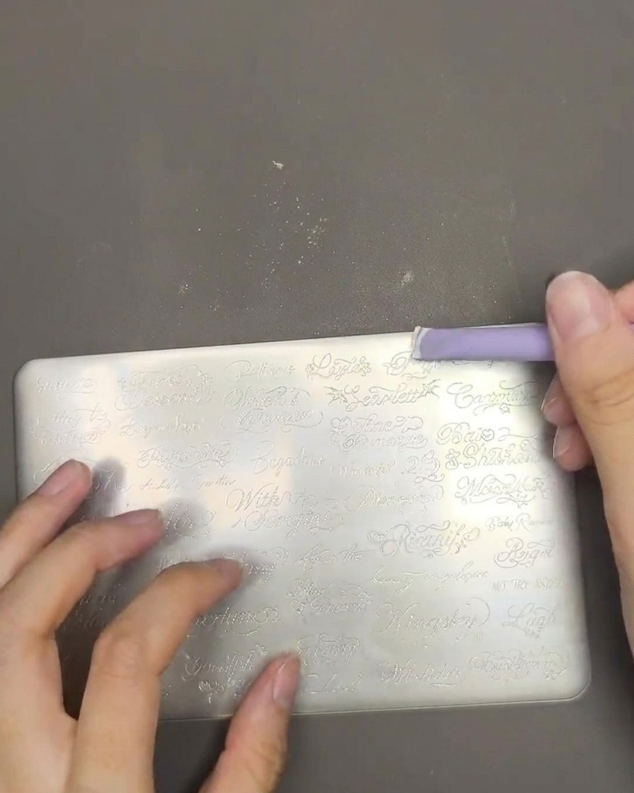 A close-up showing white stamping polish being spread across a metal stamping plate with a purple scraper.