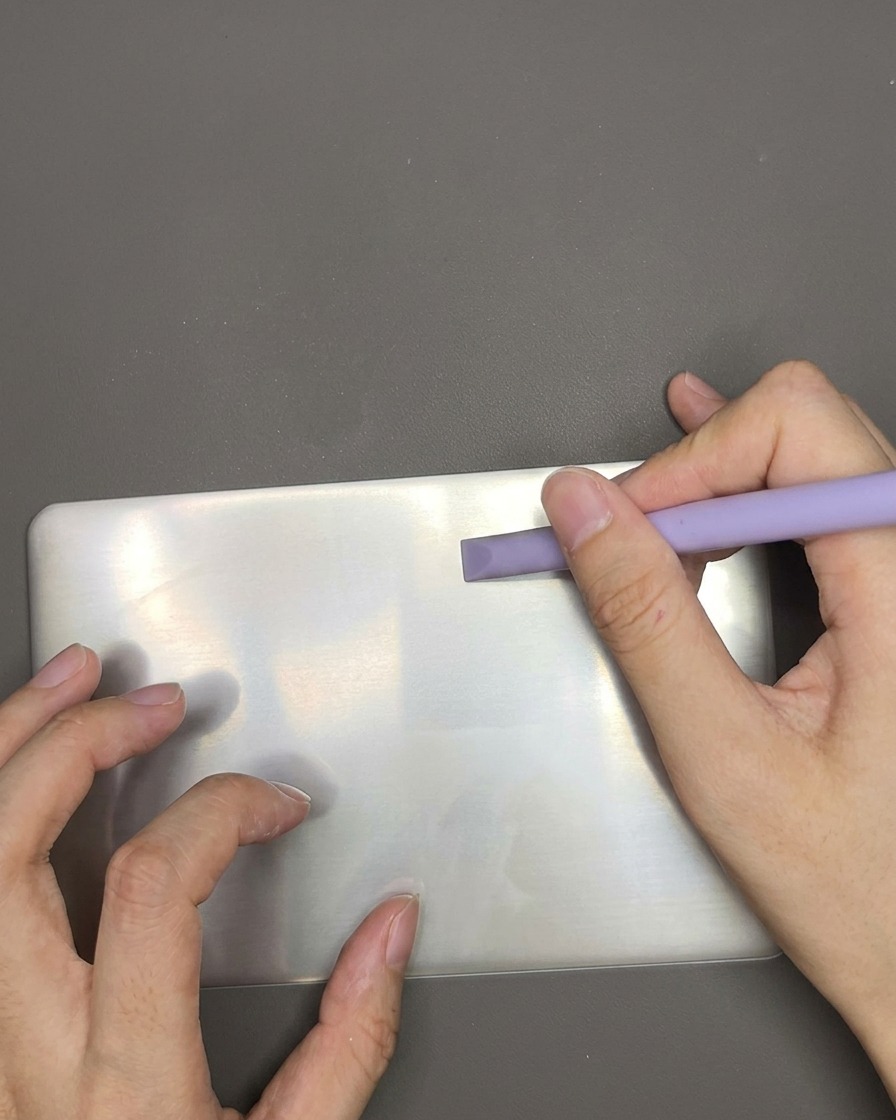 A plastic scraper tool swiping across a metal stamping plate to remove excess white polish.