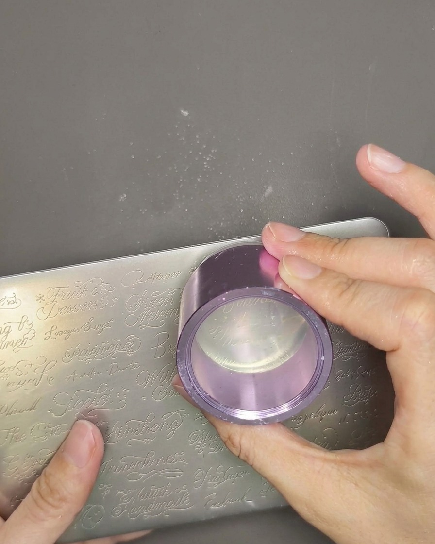 A close-up view of a clear jelly stamper head being held in hand, with a thin layer of white stamping polish applied over the previously picked-up design.