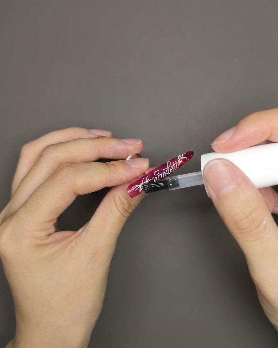 A brush applying a clear, glossy top coat over a red cat eye nail that has white English lettering stamped on it.