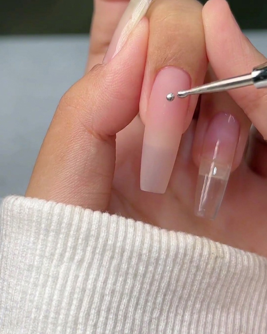 A close-up view of a dotting tool applying a single, perfect sphere of silver metallic gel onto a clean, natural nail surface.