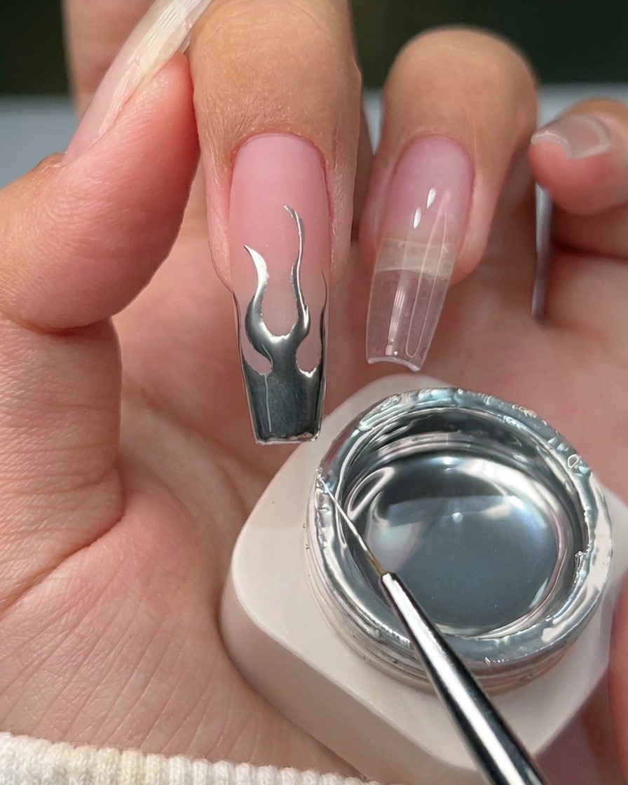 A close-up view of a finished nail with a metallic silver flame design on a neutral base.