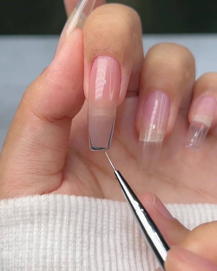 A close-up of a liner brush creating a curved silver outline on the tip of a natural nail.