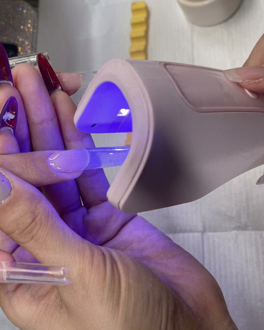 A nail artist holding a small handheld pink LED lamp over a clear square gel nail extension to cure it.