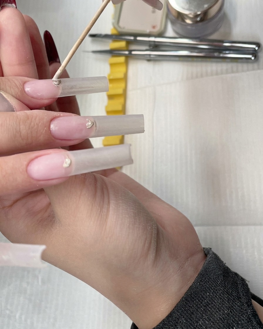A wooden stick placing a small bead of nude concealer gel onto the cuticle area of a clear nail extension.