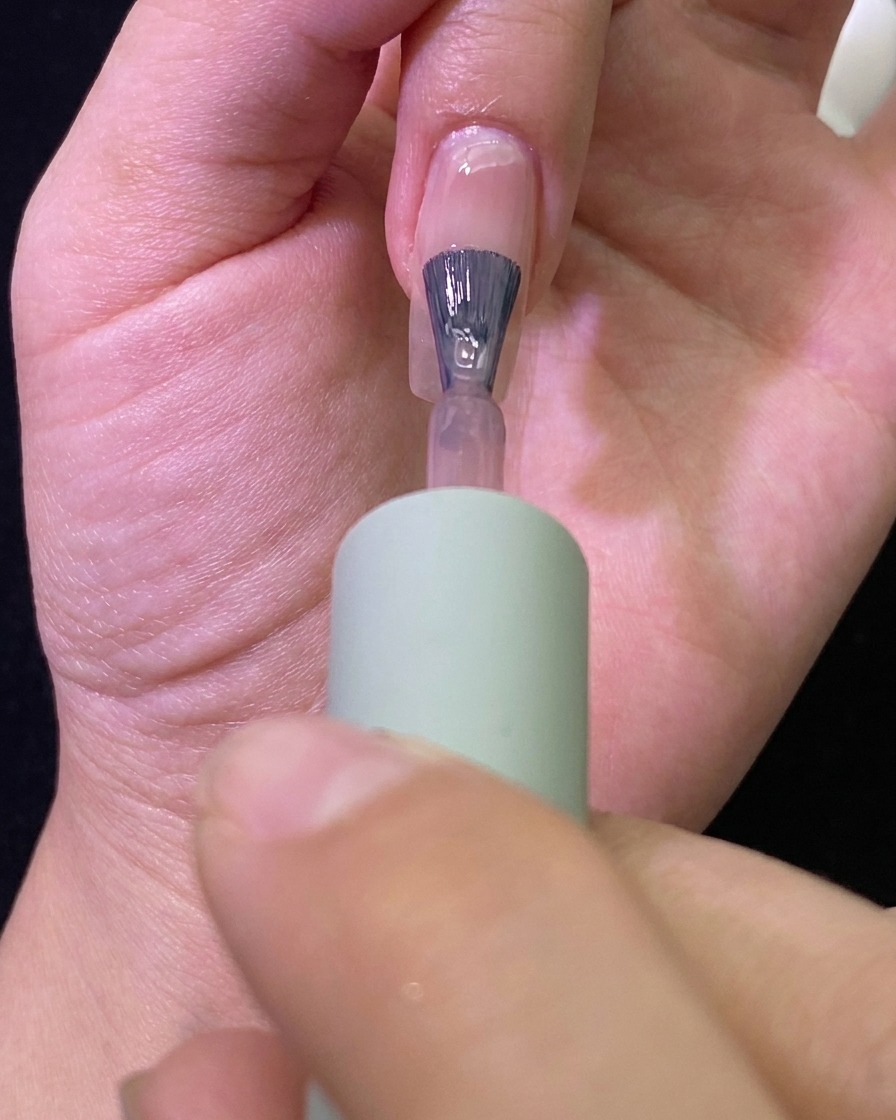 A close-up view of a brush applying a sheer nude gel polish onto a natural fingernail.