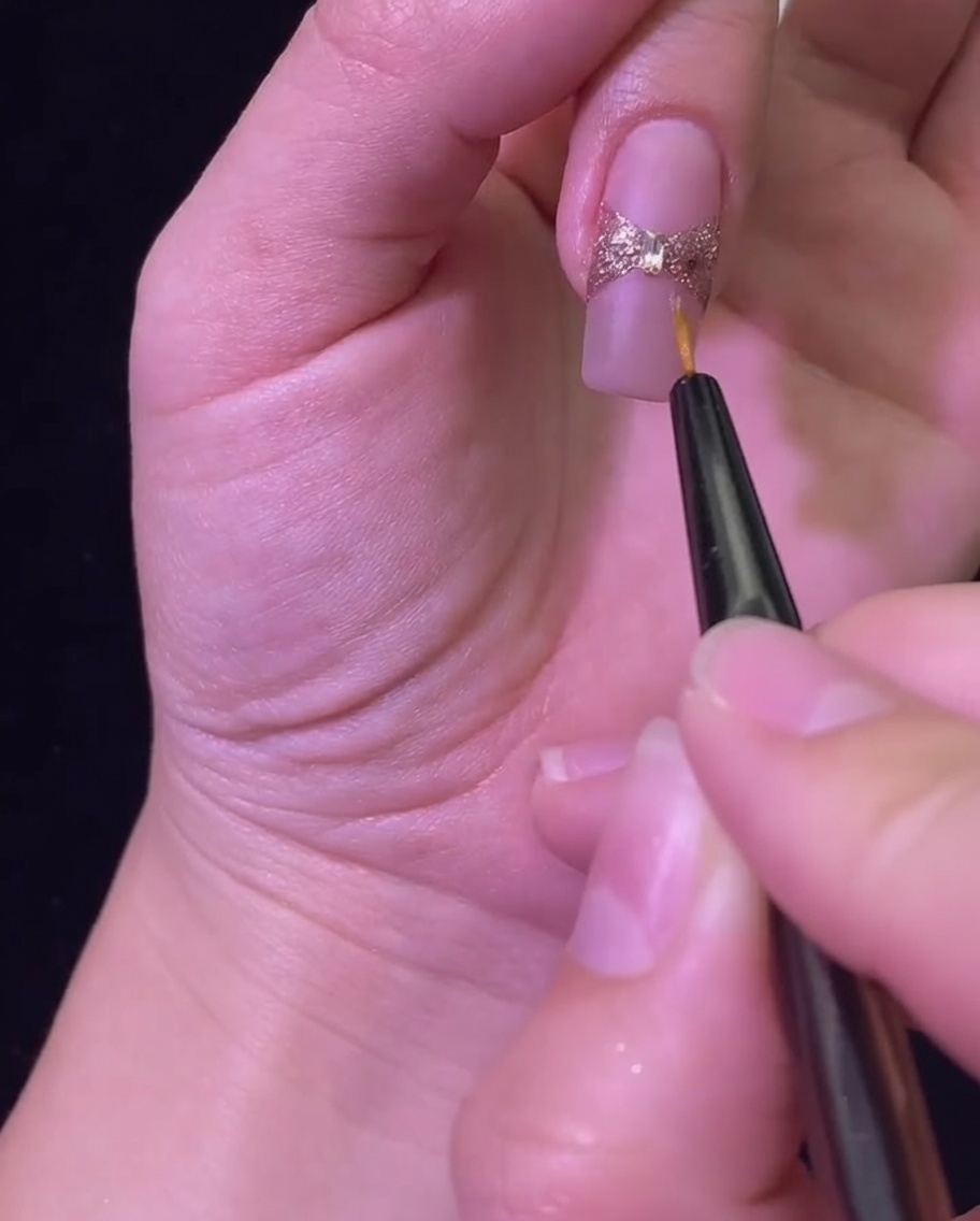 A liner brush applying a glossy top coat to seal the rose gold glitter bow and central rhinestone.